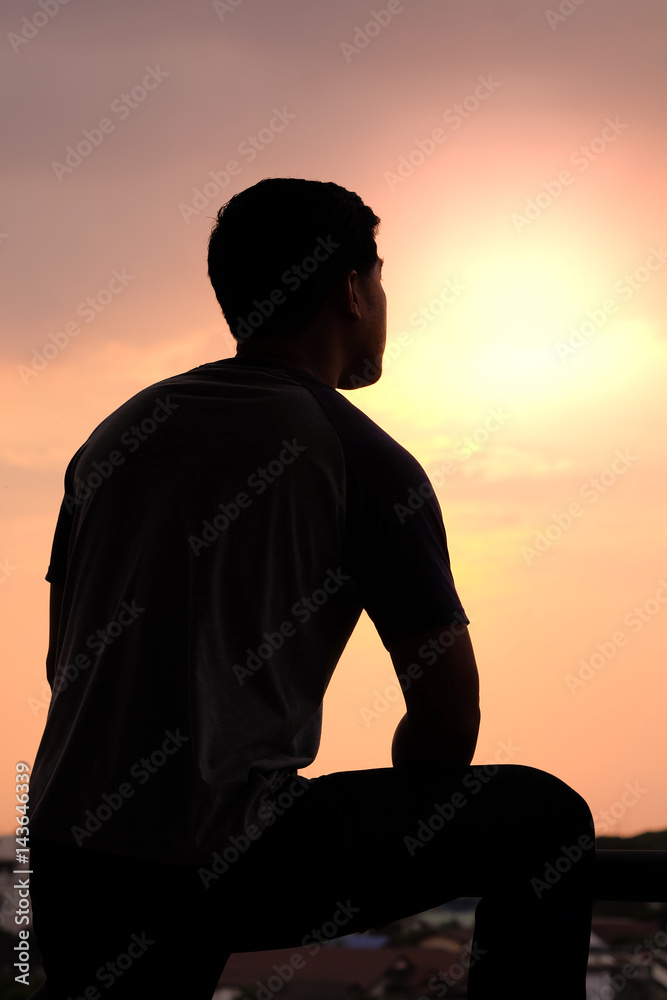 The silhouette of a man puts his arms on a leg on sunset background.