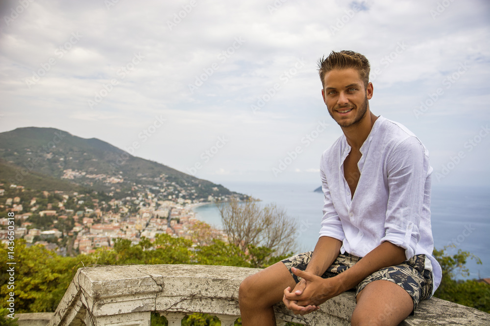 Handsome Young Man in Trendy Attire, in a Sunny Summer Day with Italian Sea Coast in the Distance, Wearing a White Shirt