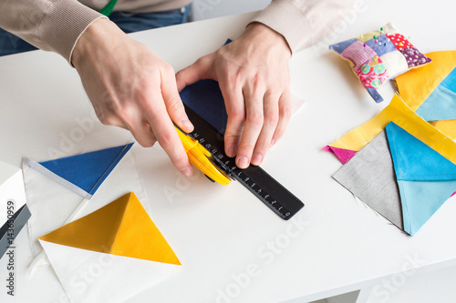 closeup on men hands of tailor, who is sitting behind a white desk and cut out colorful pieces of fabrics with rotary cutters for sewing patchwork items. Handwork and hobby concept. photo