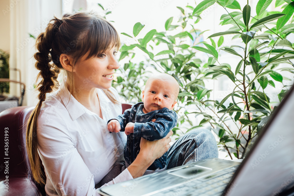 Beautiful young mother working with laptop computer and breastfeeding, holding and nursing her newborn baby at cafe. Mom - business woman feeding newborn.