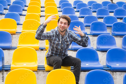 Attractive sporty young man model in blue shirt sitting on blue stadium seats after training staring at field