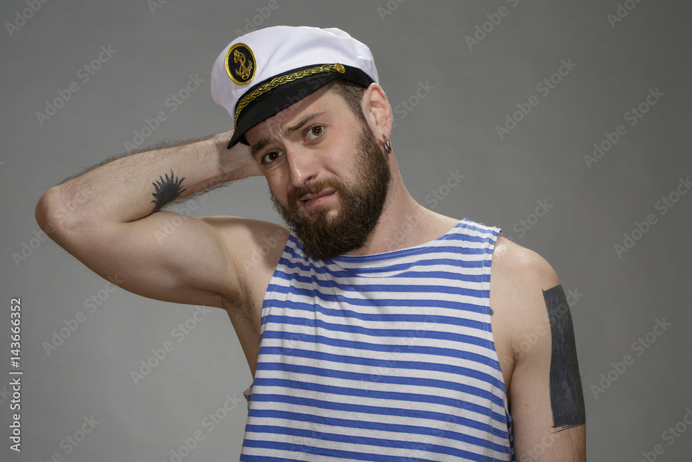  My bad. Portrait of a bearded man wearing sailor outfit looking guilty scratching the back of his head on grey background.
