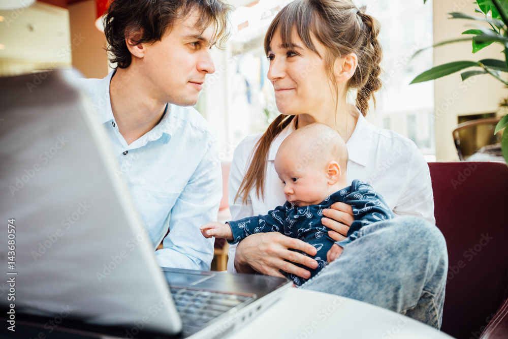 Close up uf young couple with cute little baby boy sitting on red couch with tablet computer and looking to each other in cafe interior. Close up uf young couple with cute little baby boy sitting on red couch with tablet computer and looking to each other in cafe interior.
