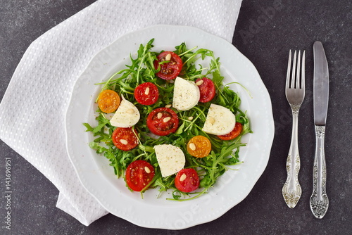 Traditional Italian salad with cherry tomato, ruccola, mozzarella, olive oil wine vinegar on a white plate on a grey abstract background.