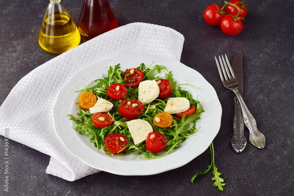 Traditional Italian salad with cherry tomato, ruccola, mozzarella, olive oil wine vinegar on a white plate on a grey abstract background.