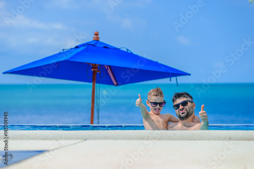 Seven years old cute boy smiling without tooth. He and his bearded father are in sunglasses, show their tongues, they leaned on pool railing giving a grimace of happiness. Sea view and beach umbrellas photo