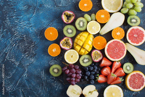Colorful raw fruits platter: oranges, lemons, kiwis, grapefruits, strawberries, grapes, blueberries, pears, passion fruit, mango on the black slate on the dark blue background, top view
