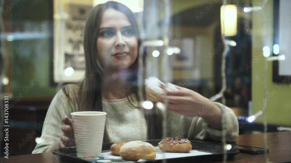 Young woman wearing a white sweater is eating a donut and drinking coffee in a cafe. Concept of a lunch break. Locked down real time medium shot Young woman wearing a white sweater is eating a donut and drinking coffee in a cafe. Concept of a lunch break. Locked down real time medium shot