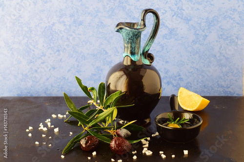 Traditional Greek ceramic olive oil jar with lemon, sea salt, olives, brunch of olive tree on a dark back ground. photo