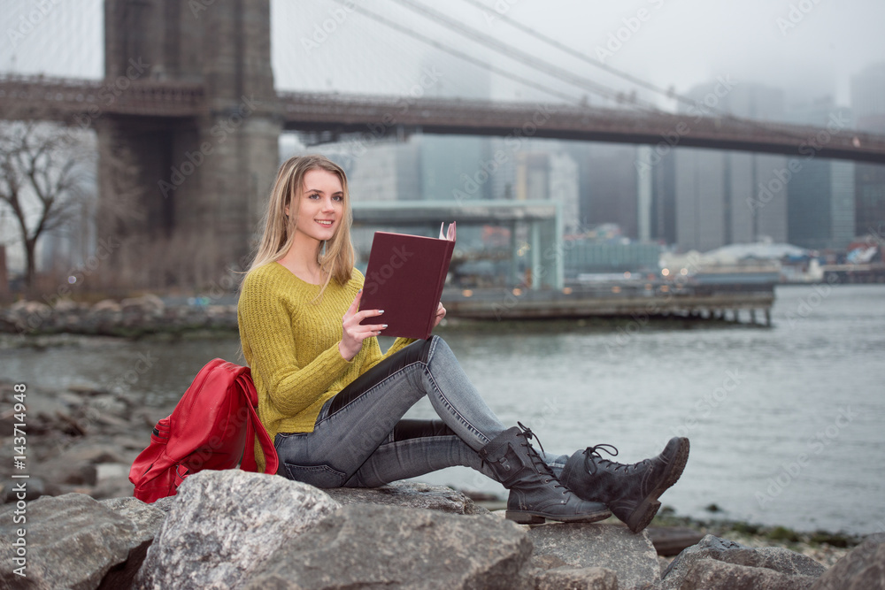 Young beautiful student girl reading a book sitting near New York City skyline. Study in USA university concept. Young beautiful student girl reading a book sitting near New York City skyline. Study in USA university concept.