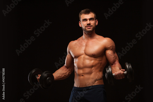 Handsome athletic man in gym is pumping up muscles with dumbbells in a gym. Fitness muscular body isolated on dark background. © satyrenko