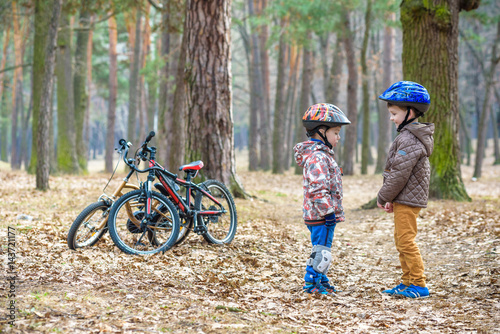 Two little kids boys, best friends in autumn forest. Older brother helping younger child to put his bike helmet. Happy siblings with bicycles.