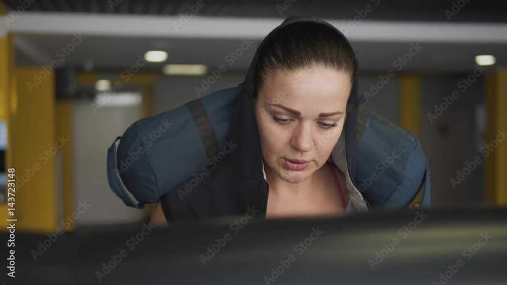 A sports woman confidently walks uphill on a treadmill with a bag of cargo in her hands. A woman trains her endurance and strength for an international fitness marathon. A sports woman confidently walks uphill on a treadmill with a bag of cargo in her hands. A woman trains her endurance and strength for an international fitness marathon.