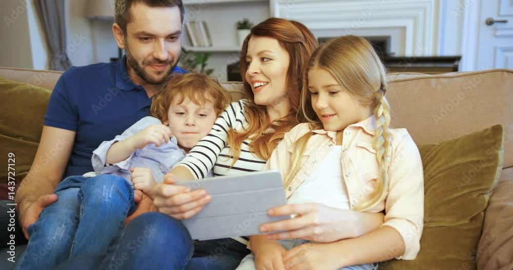 Mother, father, son and daughter using tablet, watching movie. Family spending time together at home. Happy family. Funny faces Mother, father, son and daughter using tablet, watching movie. Family spending time together at home. Happy family. Funny faces