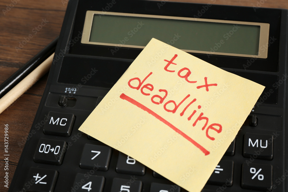 Paper sheet with text TAX DEADLINE, calculator and pencils on wooden table Paper sheet with text TAX DEADLINE, calculator and pencils on wooden table