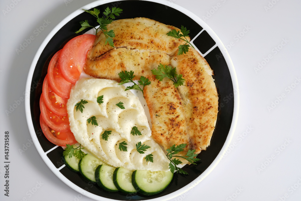 Fried tilapia fish fillets on plate with mashed potatoes, cucumber, tomatoes, parsley. 