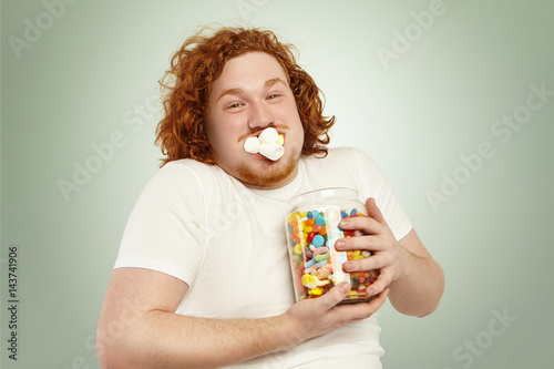 Portrait of cheerful young European overweight obese male with curly ginger hair consuming junk good, stuffing marshmallow into mouth and holding tight large glass jar, full of colorful sweets photo