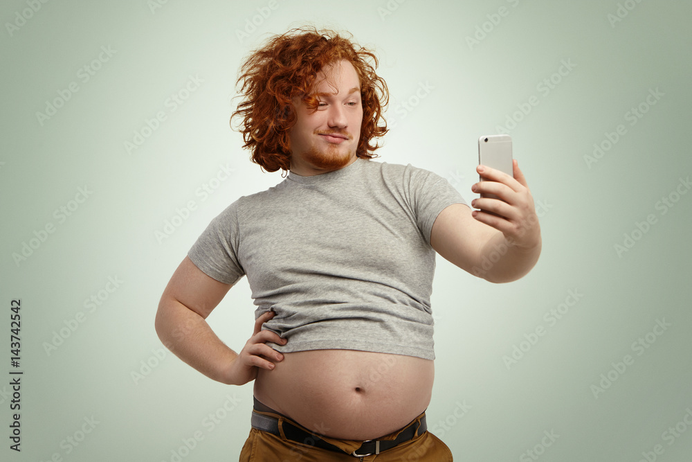 Fotografia do Stock: Funny red-haired overweight male trying to look  attractive and sexy, holding hand on his waist while taking selfie with  electronic device, belt on his pants in undone because of