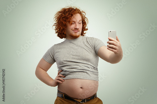 Funny red-haired overweight male trying to look attractive and sexy, holding hand on his waist while taking selfie with electronic device, belt on his pants in undone because of fat belly sticking out © wayhome.studio 