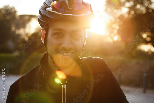 Close up shot of happy attractive young European man with beard wearing protective helmet and black sportswear looking at camera and smiling broadly during evening ride in city park on weekend © wayhome.studio 