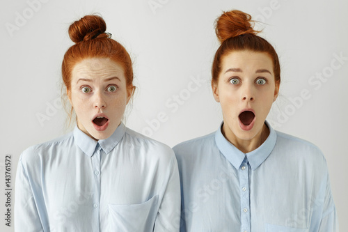 Human emotions. Two beautiful ginger girls wearing identical blue shirts and hairstyles looking in astonishment at camera with mouths widely opened and jaws dropped, surprised with shocking news photo