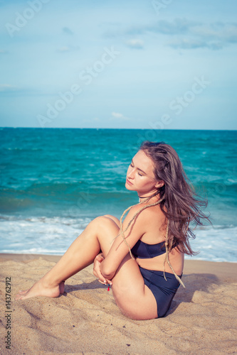 Sexy woman with tan skin in a black swimsuit sitting on the sand of the beach. Female hug her knees near sea.