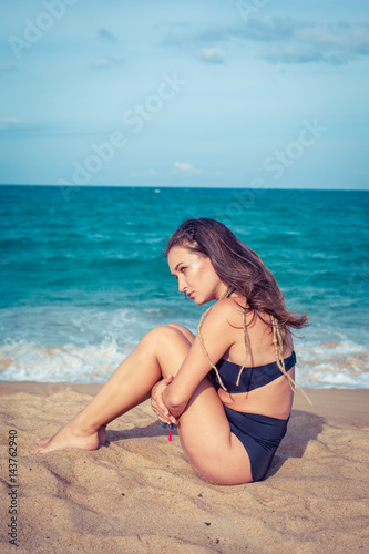Beautiful sexy woman with tan skin in a black swimsuit sitting on the sand of the beach. Female hug her knees near sea.