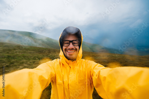 Selfie portrait of a smiling and laughing traveller man in yellow raincoat and glasses in the clouds mountains in stromy weather with rain