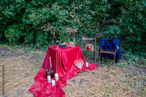 Wedding decorations outdoors. Decorated table with a tablecloth for a romantic dinner. Bottles with wax drops and white candles on a tablecloth. Heart made of wine cork. Two chairs near the table