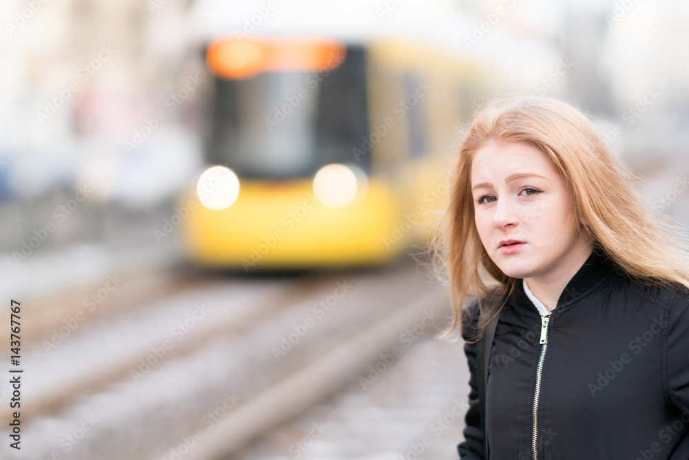 Young blonde girl posing. On background, out of focused yellow streetcar passing
