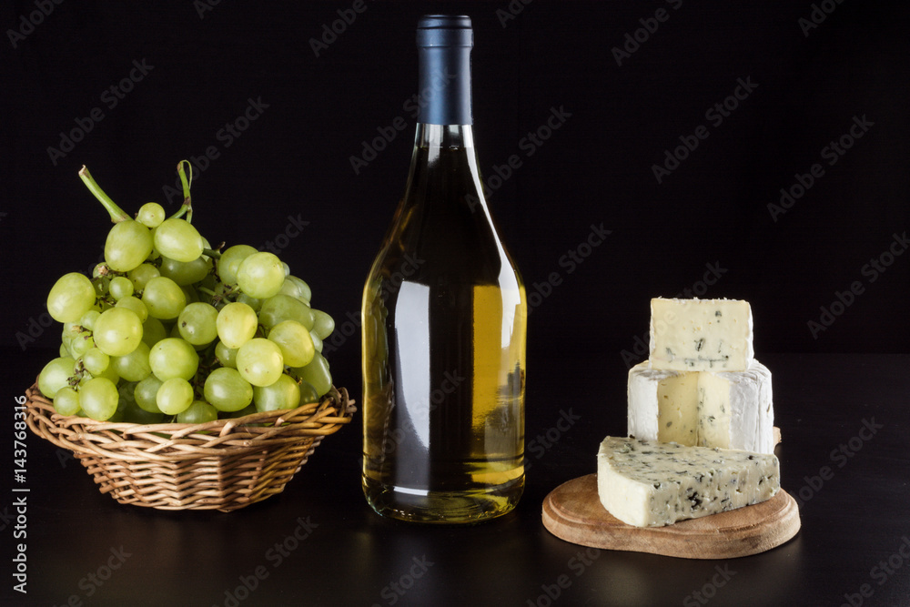 Bottle of white wine, grapes in a basket blue cheese with a mold on a wooden board on a black background, top view Bottle of white wine, grapes in a basket blue cheese with a mold on a wooden board on a black background, top view
