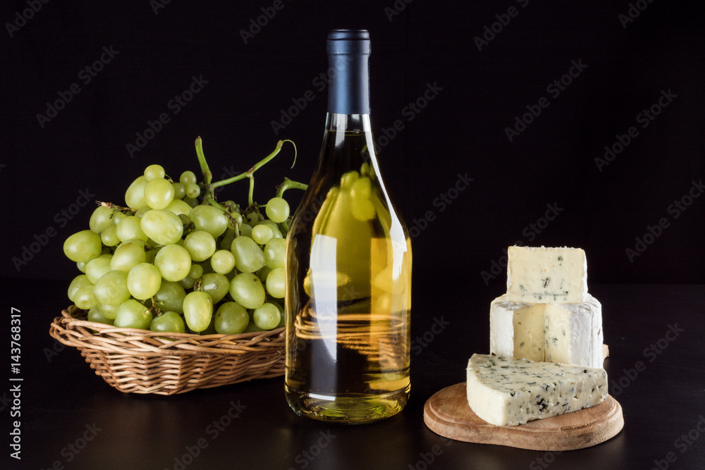Bottle of white wine, grapes in a basket blue cheese with a mold on a wooden board on a black background, top view Bottle of white wine, grapes in a basket blue cheese with a mold on a wooden board on a black background, top view