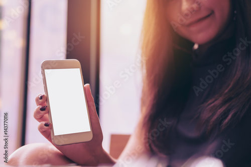 Mockup image of a beautiful woman holding and showing white mobile phone with blank white screen and smiley face in modern cafe