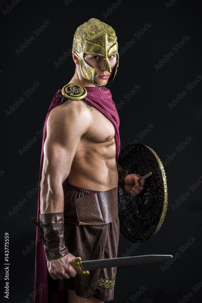 Young handsome muscular posing in roman or spartan gladiator costume shield and sword, isolated on black background in studio Stock Photo | Adobe