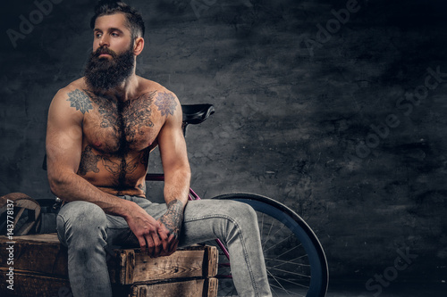 Slika na platnu Bearded male with tattooed torso sits on a wooden box.