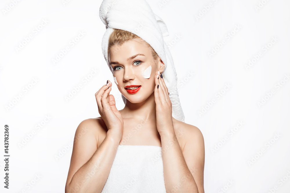 Blonde girl in a towel on her head smiling puts a moisturizing cream on her face. Portrait on a white background Blonde girl in a towel on her head smiling puts a moisturizing cream on her face. Portrait on a white background