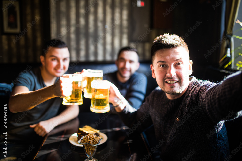 Three young men in casual clothes are smiling, taking selfie and drinking beer while sitting in pub