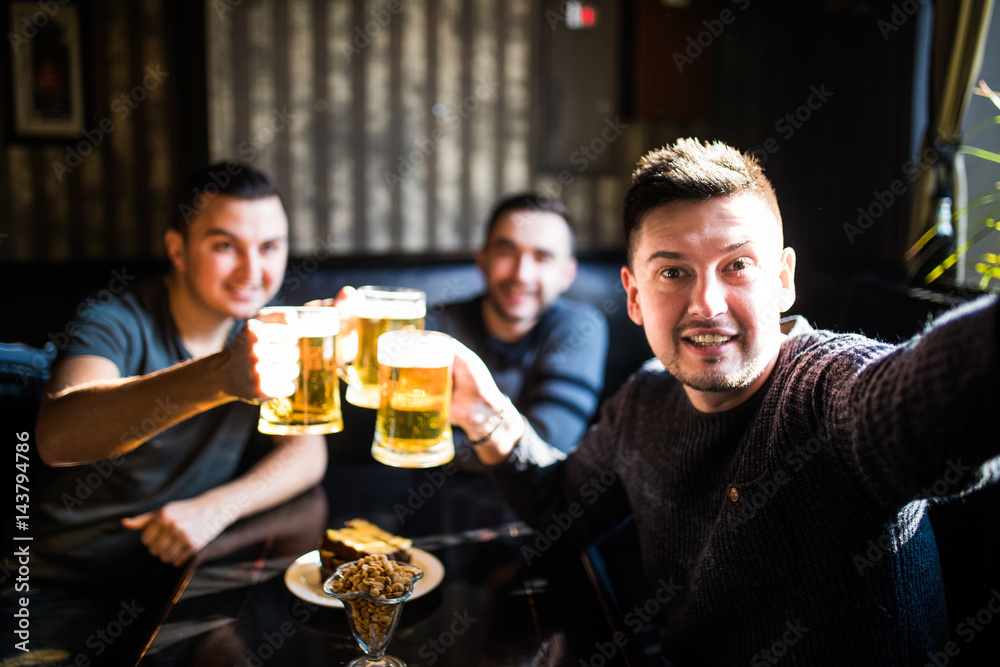 Three young men in casual clothes are smiling, taking selfie and drinking beer while sitting in pub Three young men in casual clothes are smiling, taking selfie and drinking beer while sitting in pub