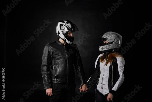 Portrait of bikers man and woman on black background. Motorcycle love concept. Extreme riders in leather jacket and suit hold hands and look at each other.