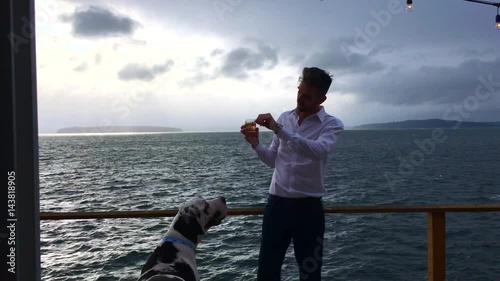 Handsome professional male drinking whiskey on seaside deck with beautiful harlequin great dane dog