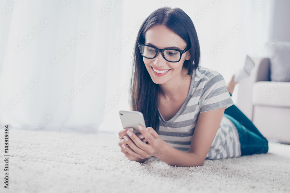 Young brunette pretty brunette girl in spectacles checking email and sms on smartphone while lying on the floor at home Young brunette pretty brunette girl in spectacles checking email and sms on smartphone while lying on the floor at home