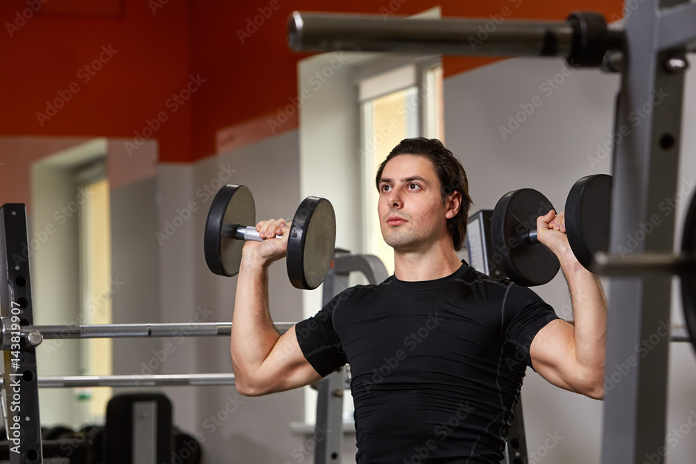 Young muscular built athlete in black sportwear sitting on a weightlifting machine and lifting two dumbbells Young muscular built athlete in black sportwear sitting on a weightlifting machine and lifting two dumbbells
