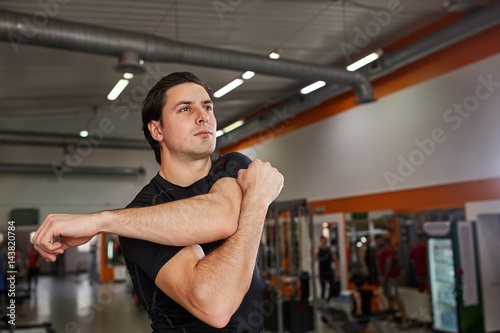 Full-length of young man in black sportswear stretching his arm at gym.
