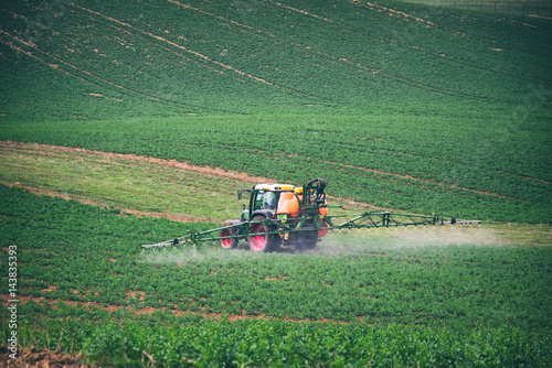 Farm machinery spraying insecticide to the green field, agricultural natural seasonal spring background. Tractor sprinkling pesticides against bugs on agricultural field