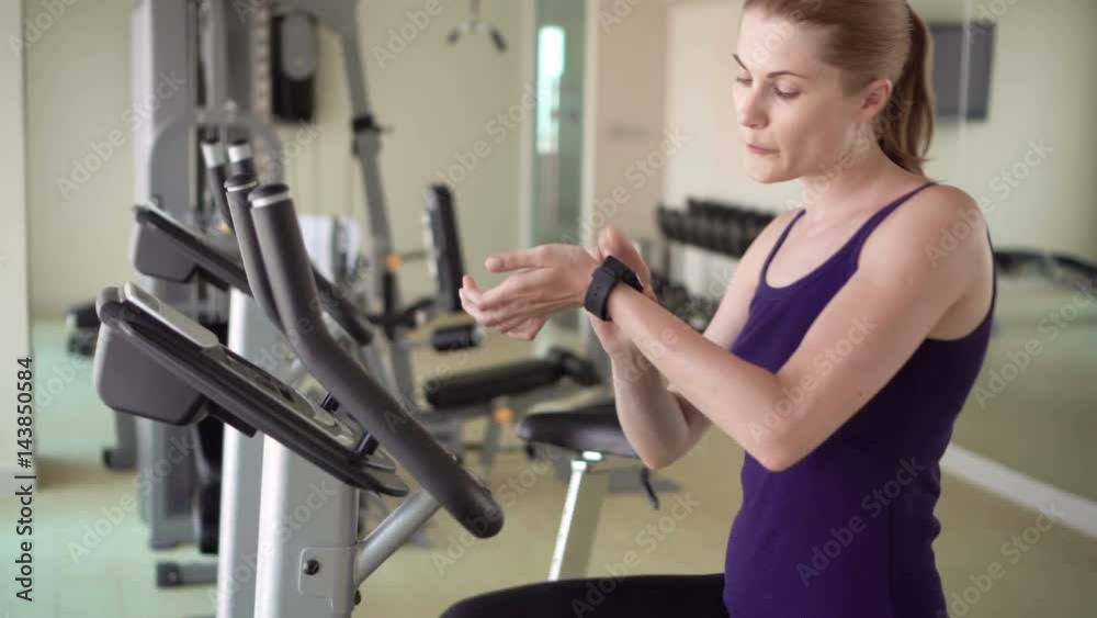 Beautiful fit young woman in gym doing exercises on a velosimulator. Using her smartwatch, browsing Beautiful fit young woman in gym doing exercises on a velosimulator. Using her smartwatch, browsing