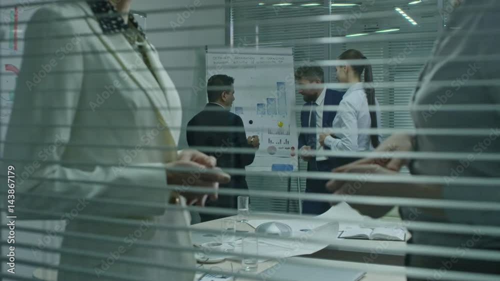 Mid-section shot through window of conference room of businesswomen gesturing and discussing work; colleagues talking about financial charts in background Mid-section shot through window of conference room of businesswomen gesturing and discussing work; colleagues talking about financial charts in background