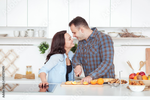 Couple making fresh organic juice in kitchen together. A young man slices a baguette. A woman standing near her boyfriend. Whiite kitchen with daylight. Healthy lifestyle concept