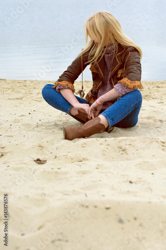 A blonde girl dressed stylishly in an American style is resting on the beach on a cloudy day, near the water she throws about her life, thinks and gets a good day have a beautiful look