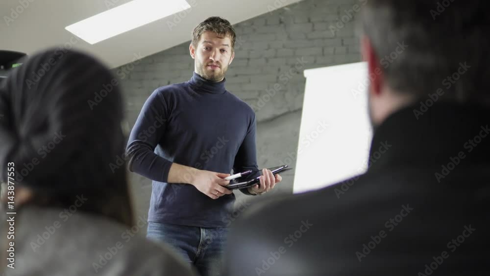 Handsome young businessman pointing at flipchart during presentation in conference room and holding tablet. Presentation speech with flipchart in office. Shot in 4k Handsome young businessman pointing at flipchart during presentation in conference room and holding tablet. Presentation speech with flipchart in office. Shot in 4k