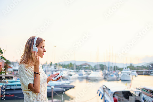 Enjoying the sound. Happy young woman with earphones is listening music on broadwalk in marine harbor.